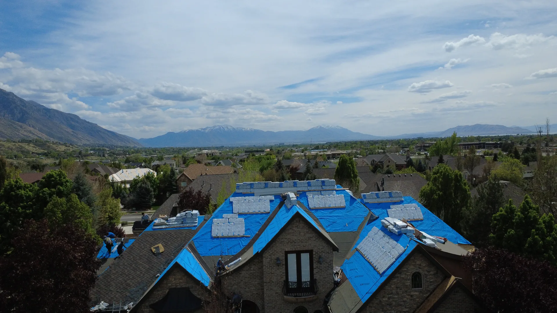 Aerial drone photograph of a Heber Valley luxury custom home reroof in progress by Frame Roofing Utah, with blue synthetic underlayment fully installed, shingle bundles staged across the roof planes, and the Wasatch Back mountain range visible in the background under a blue sky