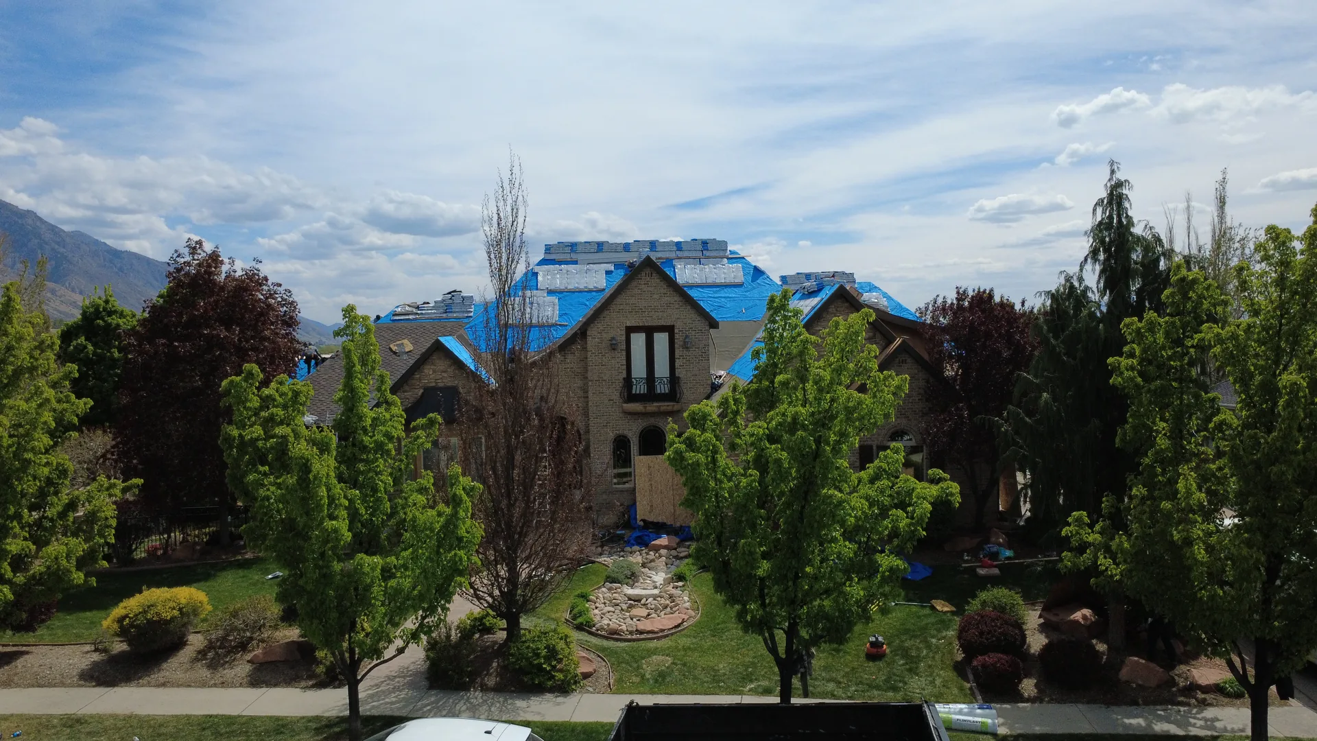 Drone photograph of the same Heber Valley custom home reroof taken from the front yard elevation, showing the two-story brick-and-stone elevation with the blue synthetic underlayment visible on the roof and the Frame Roofing Utah roll-off dumpster on the driveway.